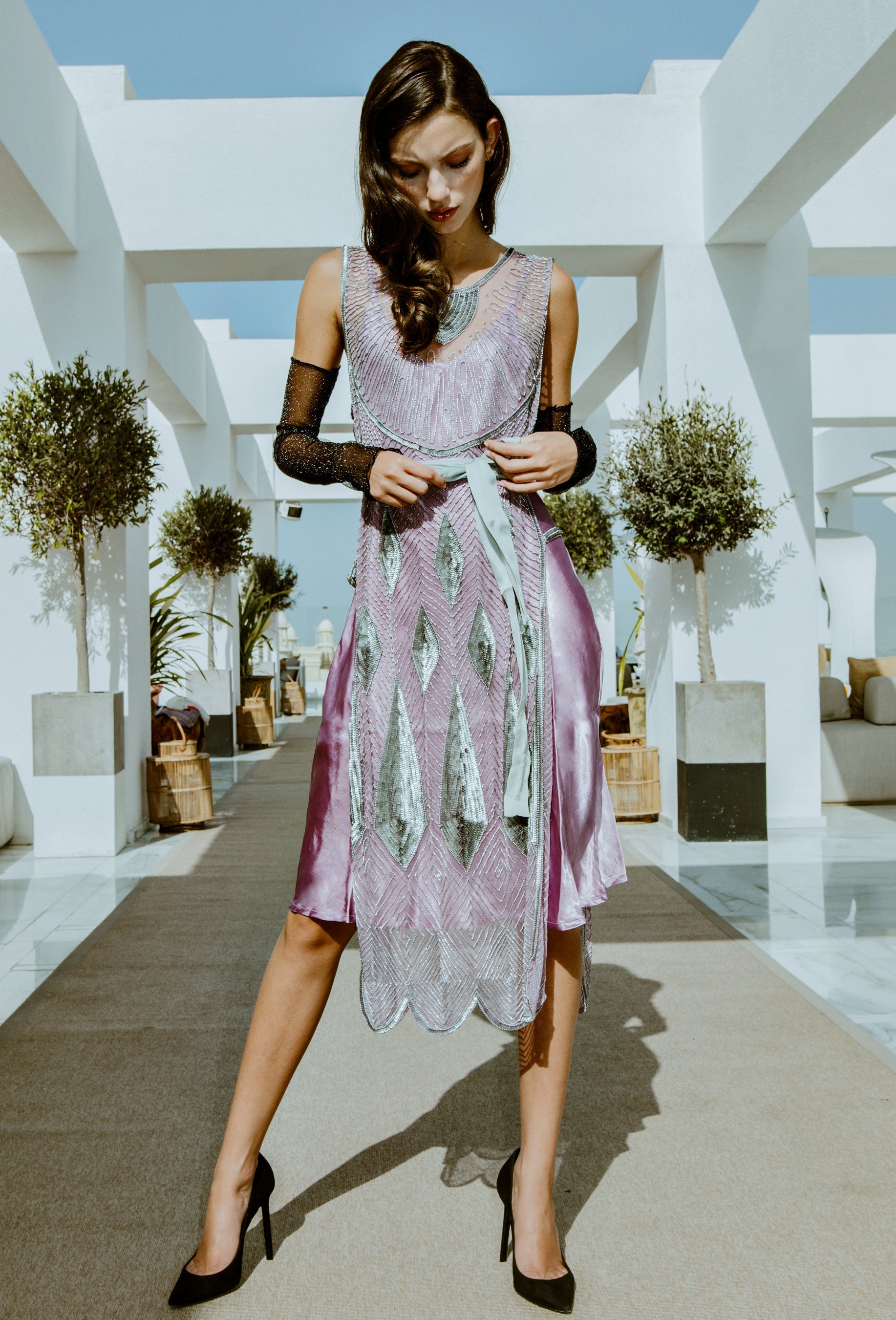 A woman models a sleeveless lavender and silver patterned midi dress with sheer black lace sleeves, styled with black pointed-toe heels, set against a modern white architectural background.