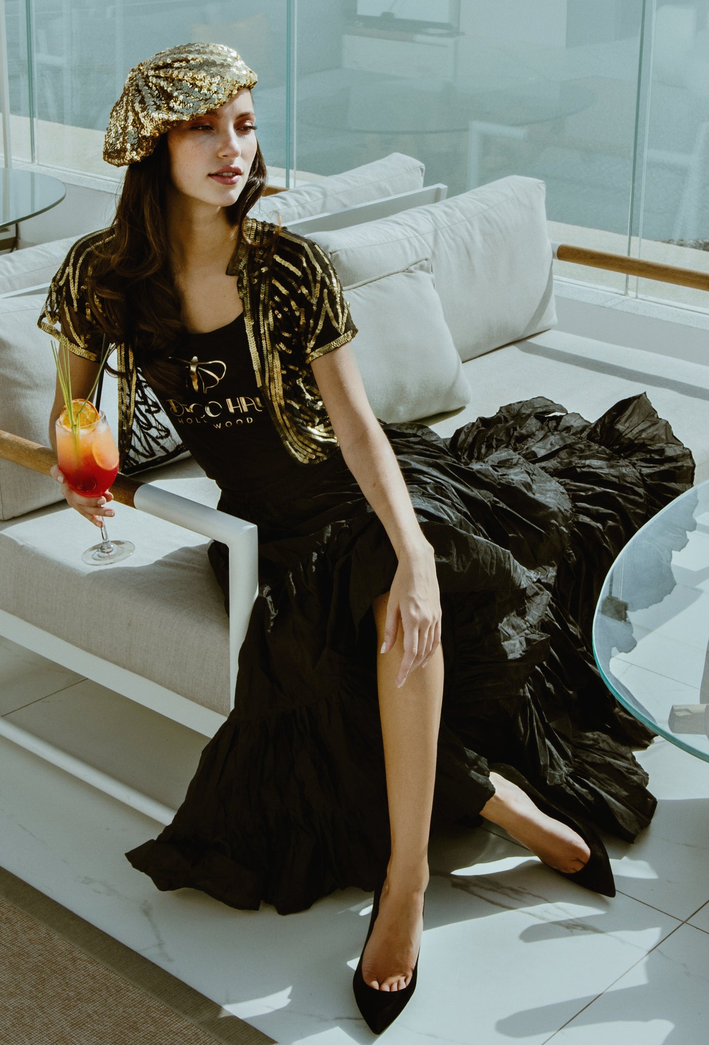 A woman models a black evening gown with gold metallic embroidery and a matching gold patterned hat, sitting on a modern white lounge sofa.